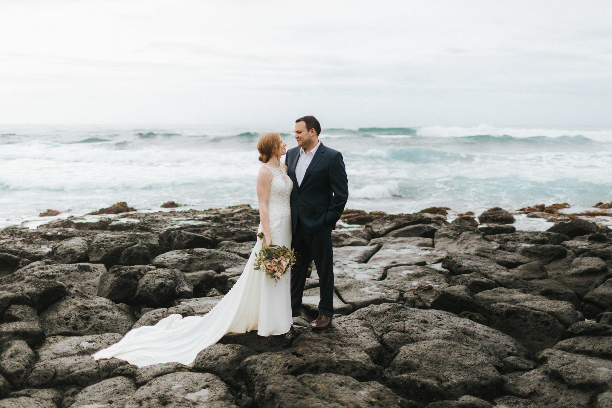 Bride and groom embrace on rocky shore on Kauai, Hawaii