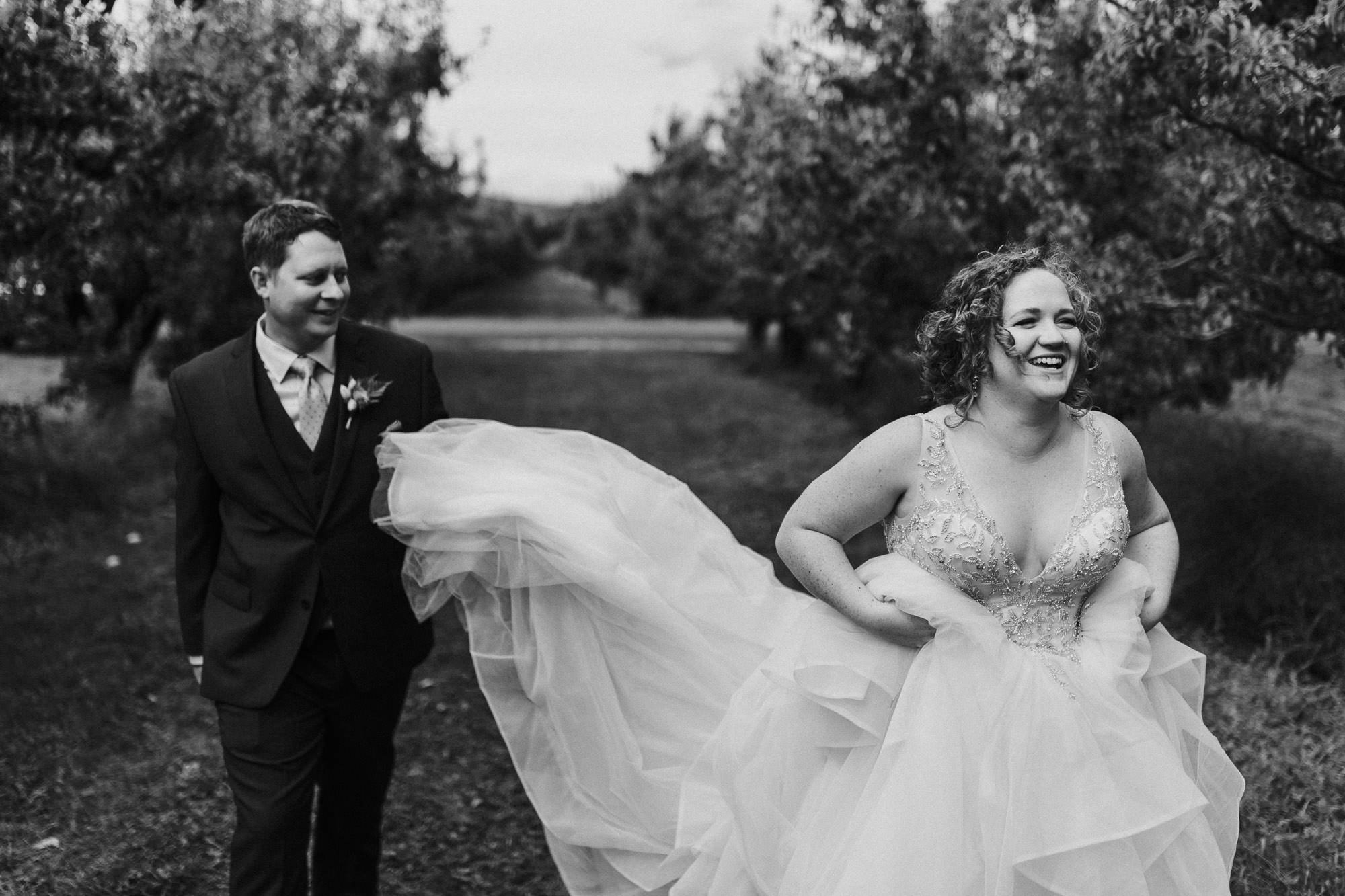 Groom holds bride's dress train as they walk through an orchard in Hood River, Oregon