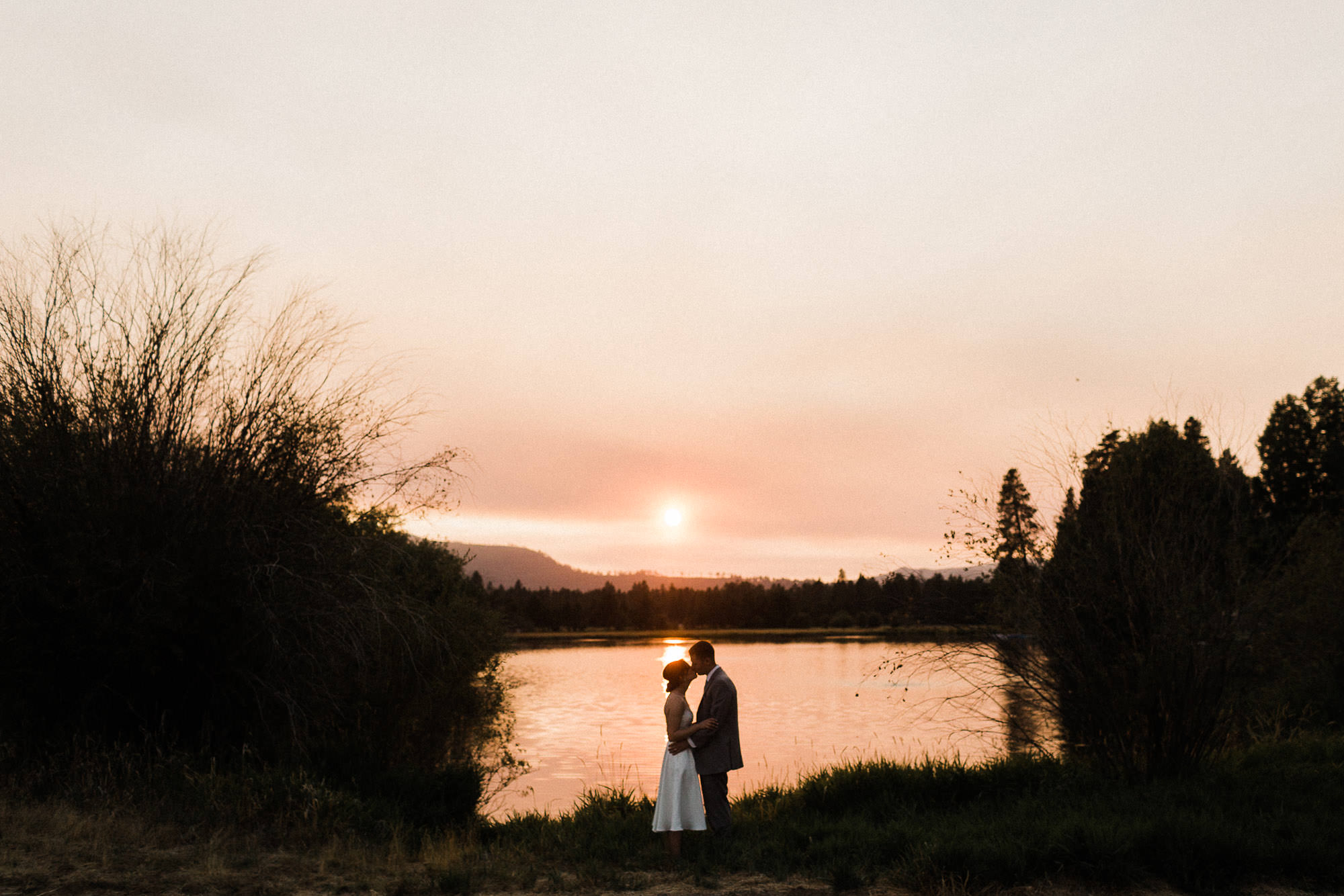 Bride and groom are silhouetted as they kiss in front of a pond at sunset at Black Butte Ranch