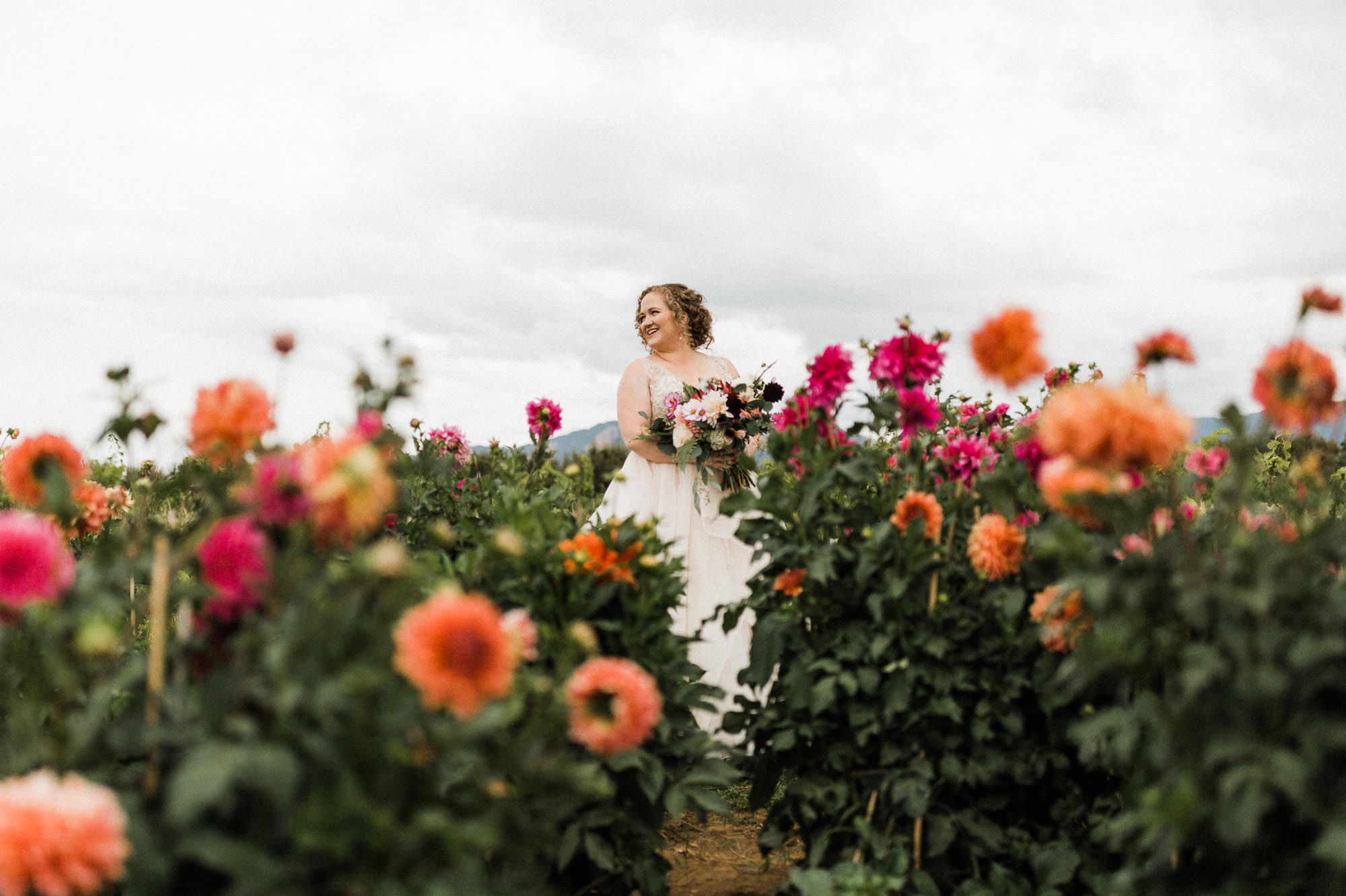The bride poses for a portrait in a field of orange and pink dahlias in Mt. Hood, Oregon.