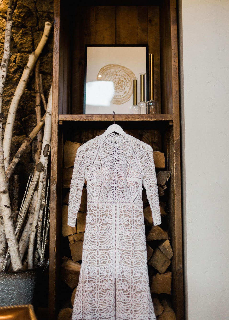 A stunning white, sleeved wedding dress hung on a wooden bookcase. Sitting inside the bookcase are large pieces of firewood. 