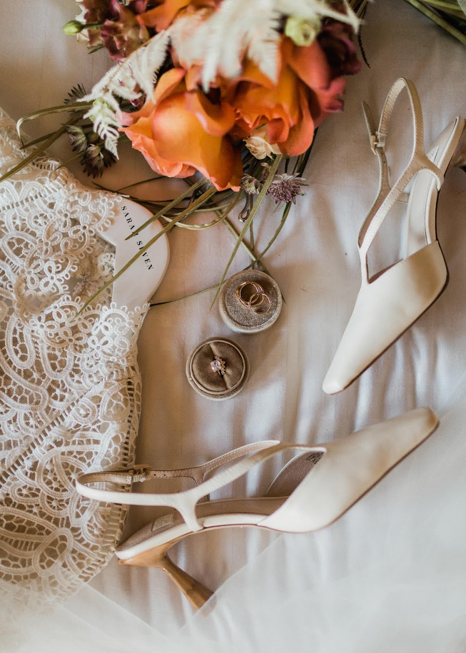A detailed shot of the bridal accessories including closed-toed heels, diamond wedding ring along with gold wedding bands, colorful florals and a portion of the wedding dress. 