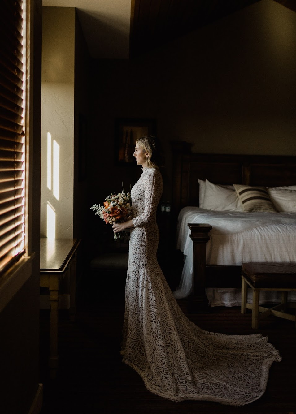 The bride is in her room, staring out the window. The train from her dress is laid out behind her as she holds her bouquet.