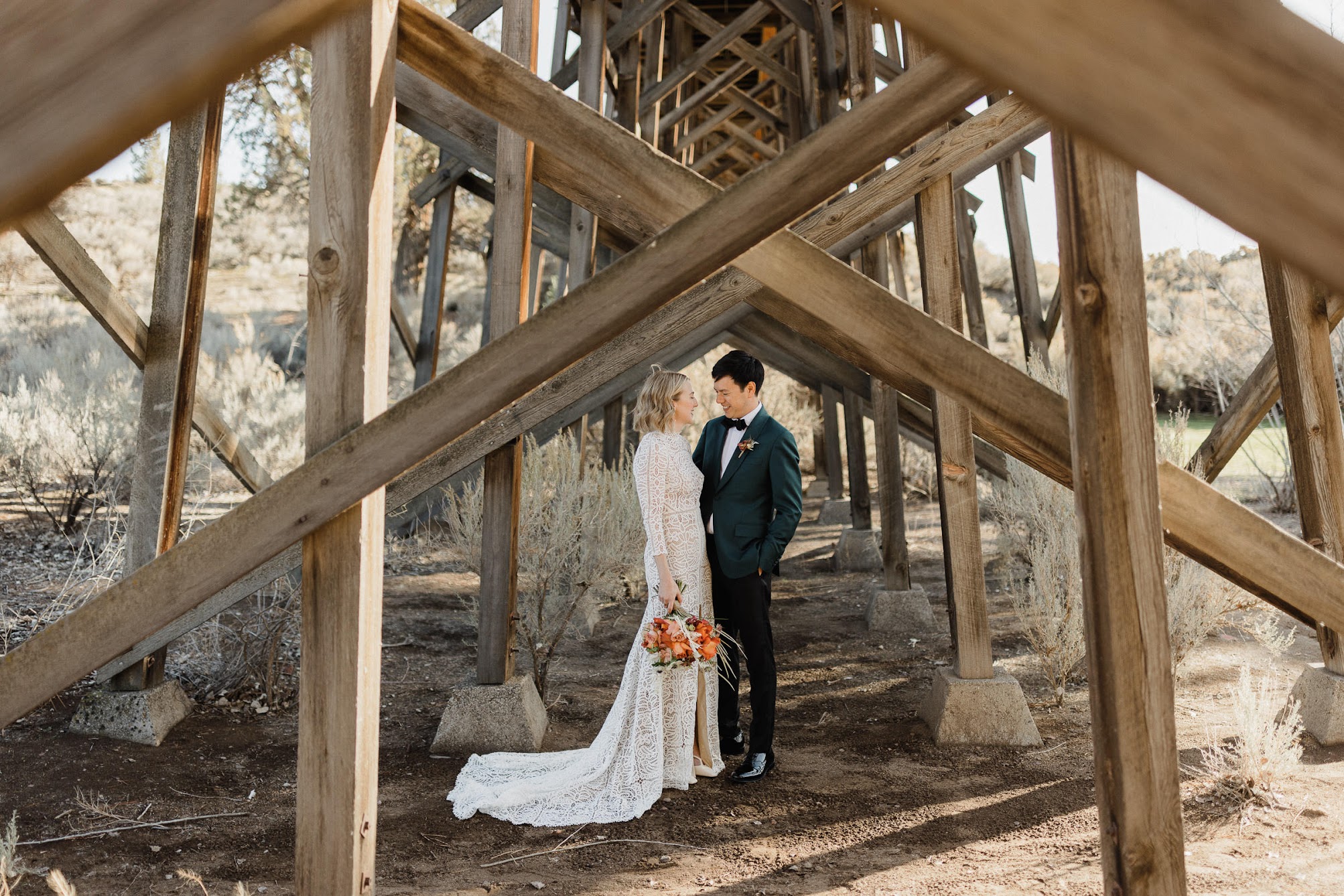 Bride and groom standing under a rustic bridge, looking into each other's eyes. 