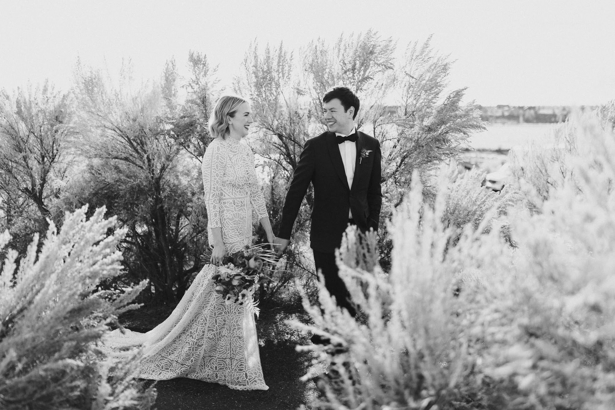 Bride and groom walking through dried bushes while holding hands looking at each other.
