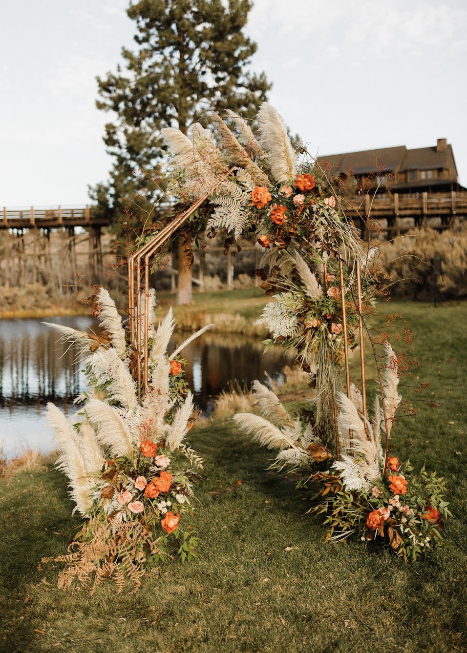 brass arbor adorned with wispy pampas grass, dried ferns, and warm florals. Set in front of a small pond, with a rustic bridge and barn in the background.