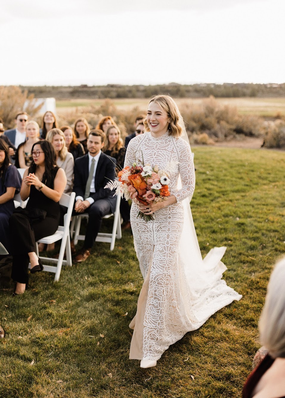 The bride is walking down the grassy aisle alone with her bouquet in her hand smiling. 