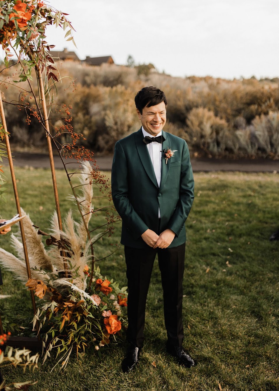 The groom is standing at the arbor, hands folded in front of him while he smiles. 