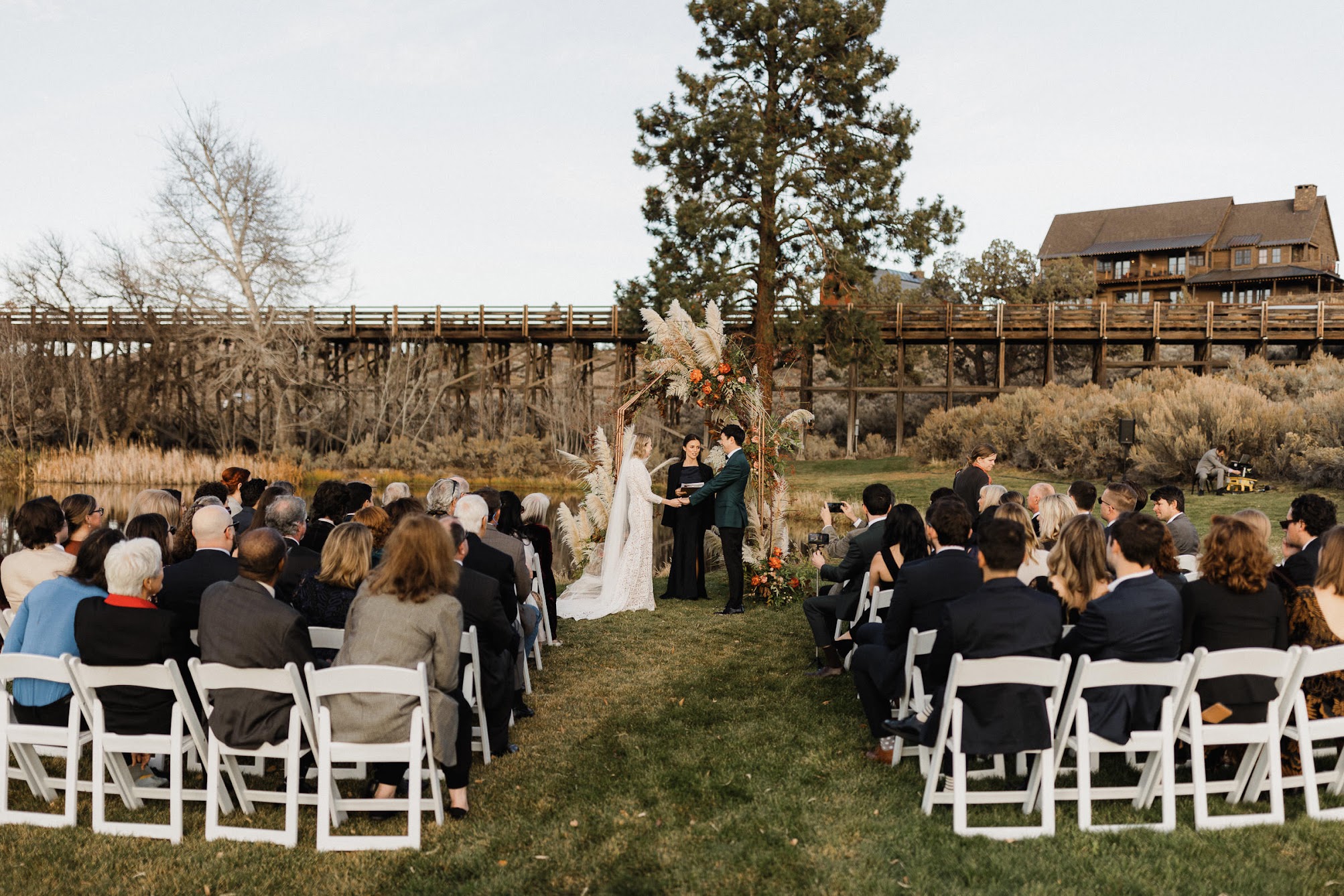 The bride and groom holding hands at the arbor while the officiant stands in front of them. Their guests are all sitting down watching them. 