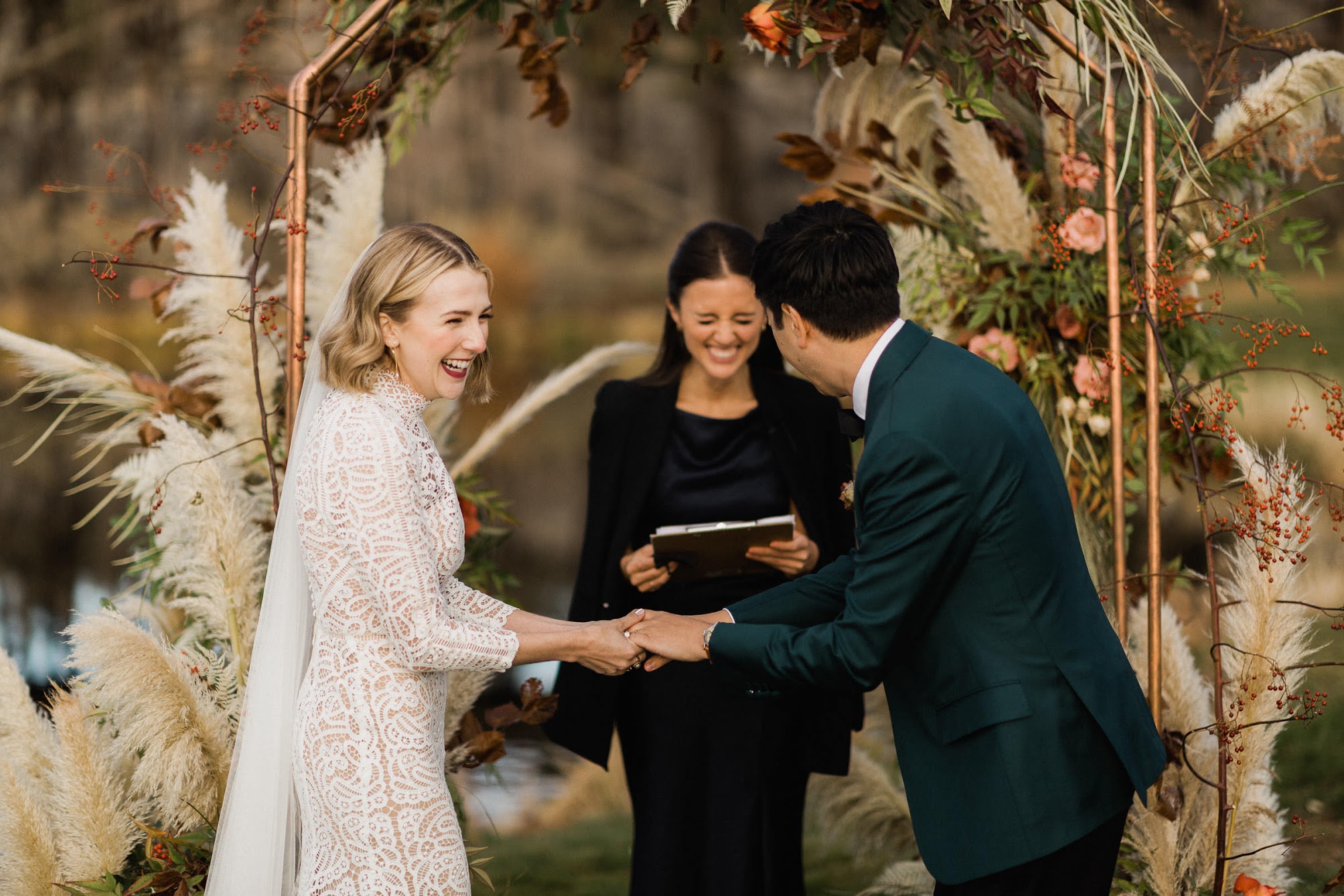The bride, groom and officiant sharing a laugh at the arbor while the bride and groom hold hands.