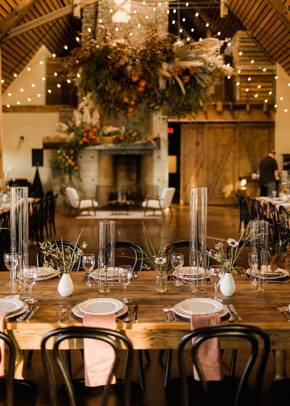 Farm tables are set with velvet pink napkins draped over the table sitting between the dinner and salad plate. Tall black taper candles with a glass chimney around them, in between each candles sits a small bud vase.