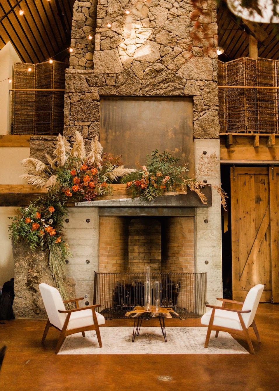 a large brick fireplace covered in tons of florals and pampas grass. In front of the fireplace are two modern-looking chairs, and a coffee table made out of the base of a tree with tapered candles on it.
