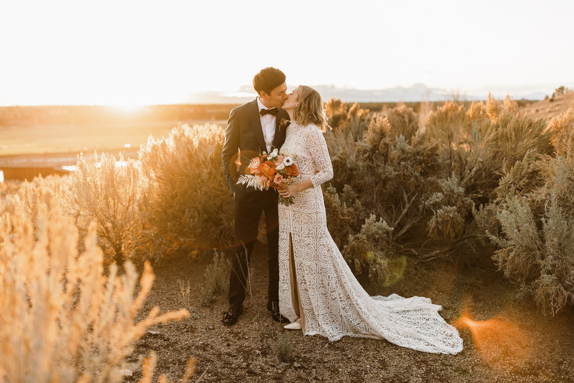 Stunning sunset photos. The bride and groom are kissing in a field surrounded by dried bushes. Behind them is open farm land and the sunsetting.  