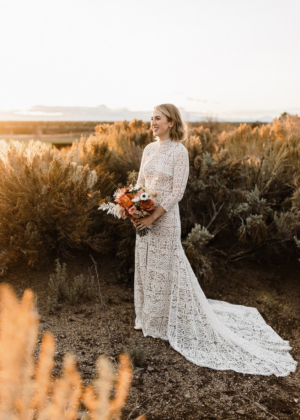 the bride staring out into the field with a big smile on, as she hold her floral bouquet and the sun is setting behind her. 