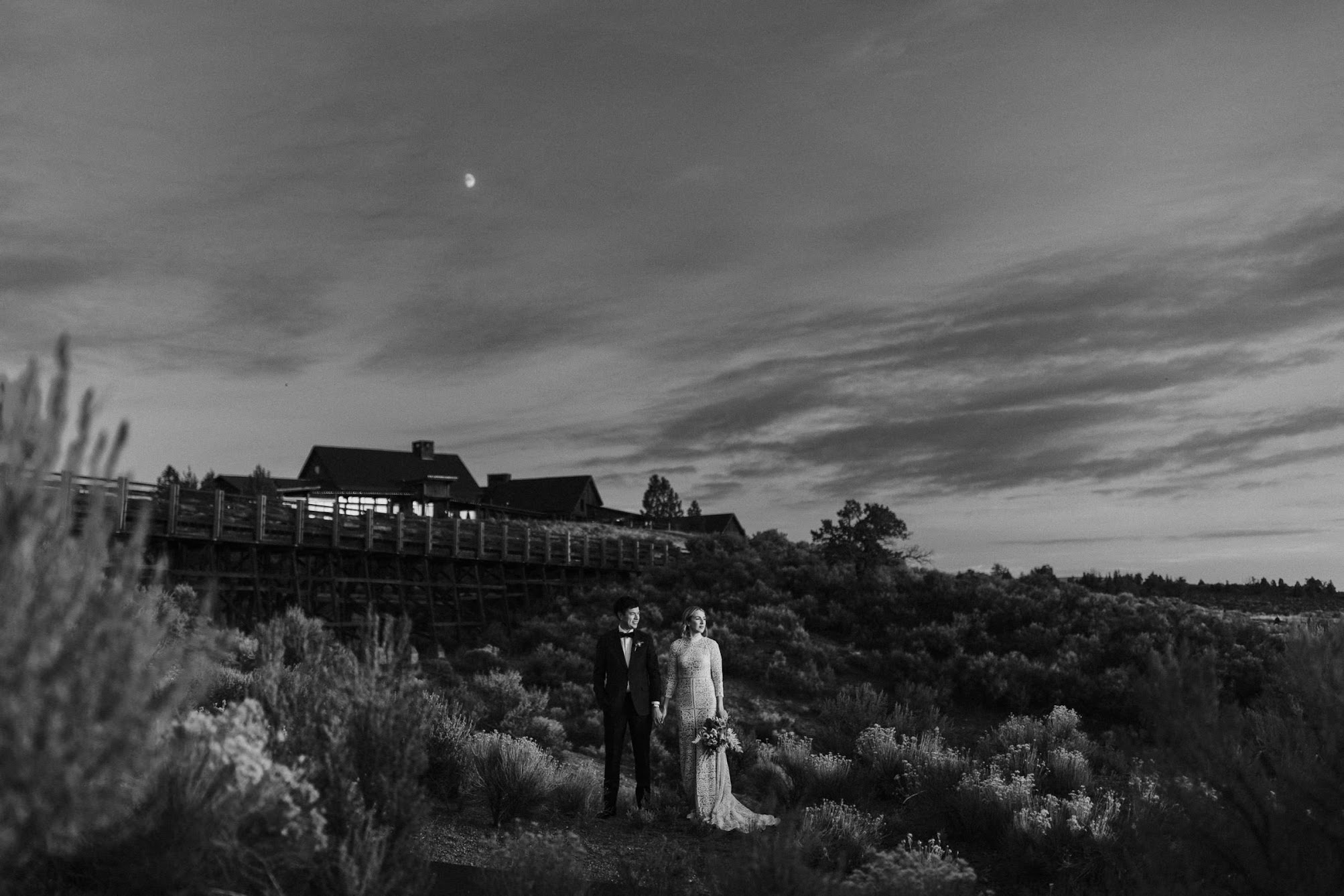 A stunning black and white photo, the bride and groom on holding hands staring into the distance. While Brasada Ranch and the rustic wood bride sit behind them. Above the couple is half a moon.
