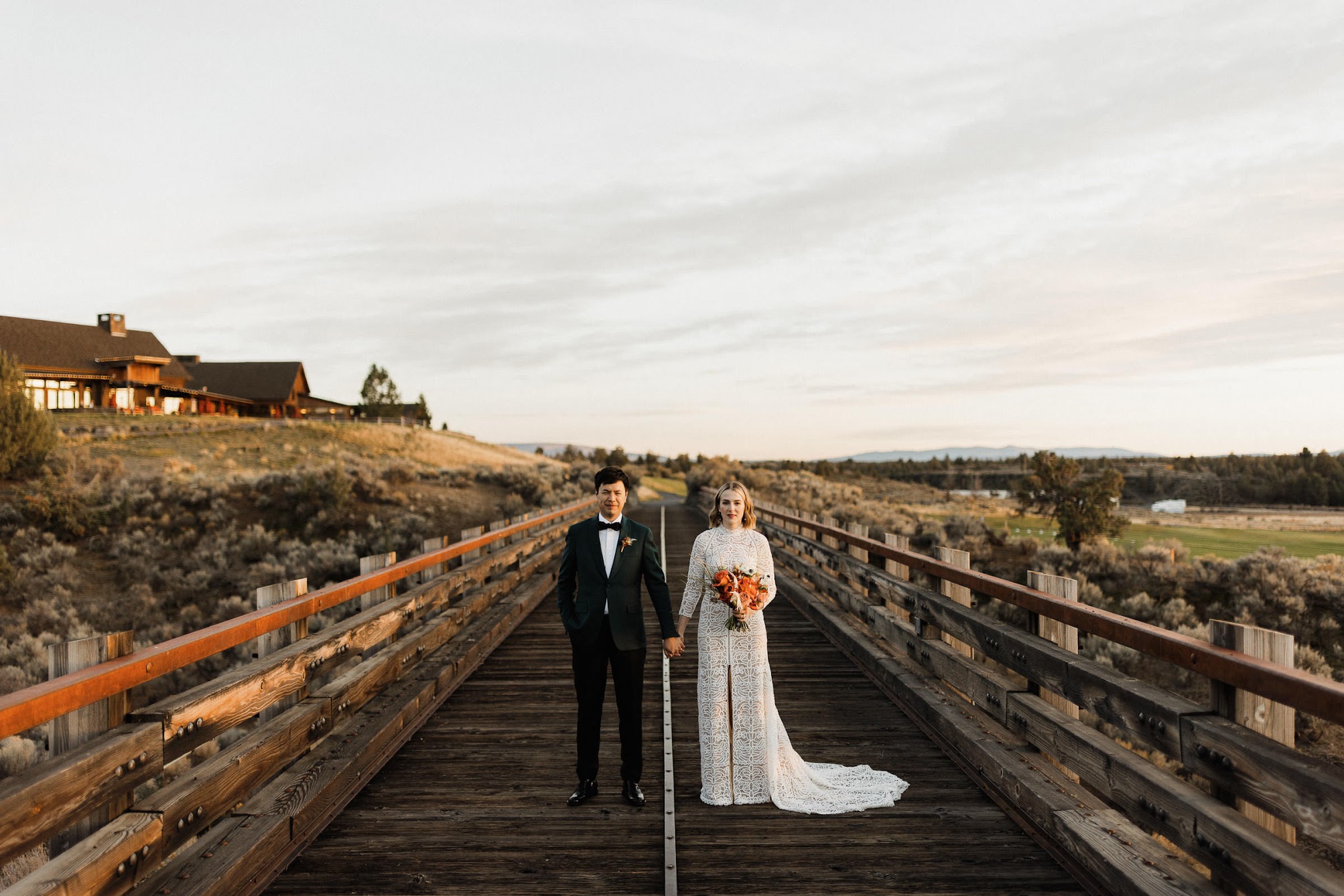 The bride and groom standing on a long wooden bridge holding hands with a serious faces on