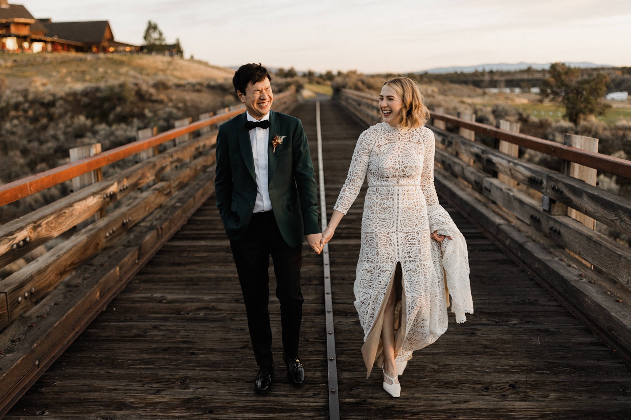 The bride and groom walk along a long, wooden, rustic bridge laughing. 