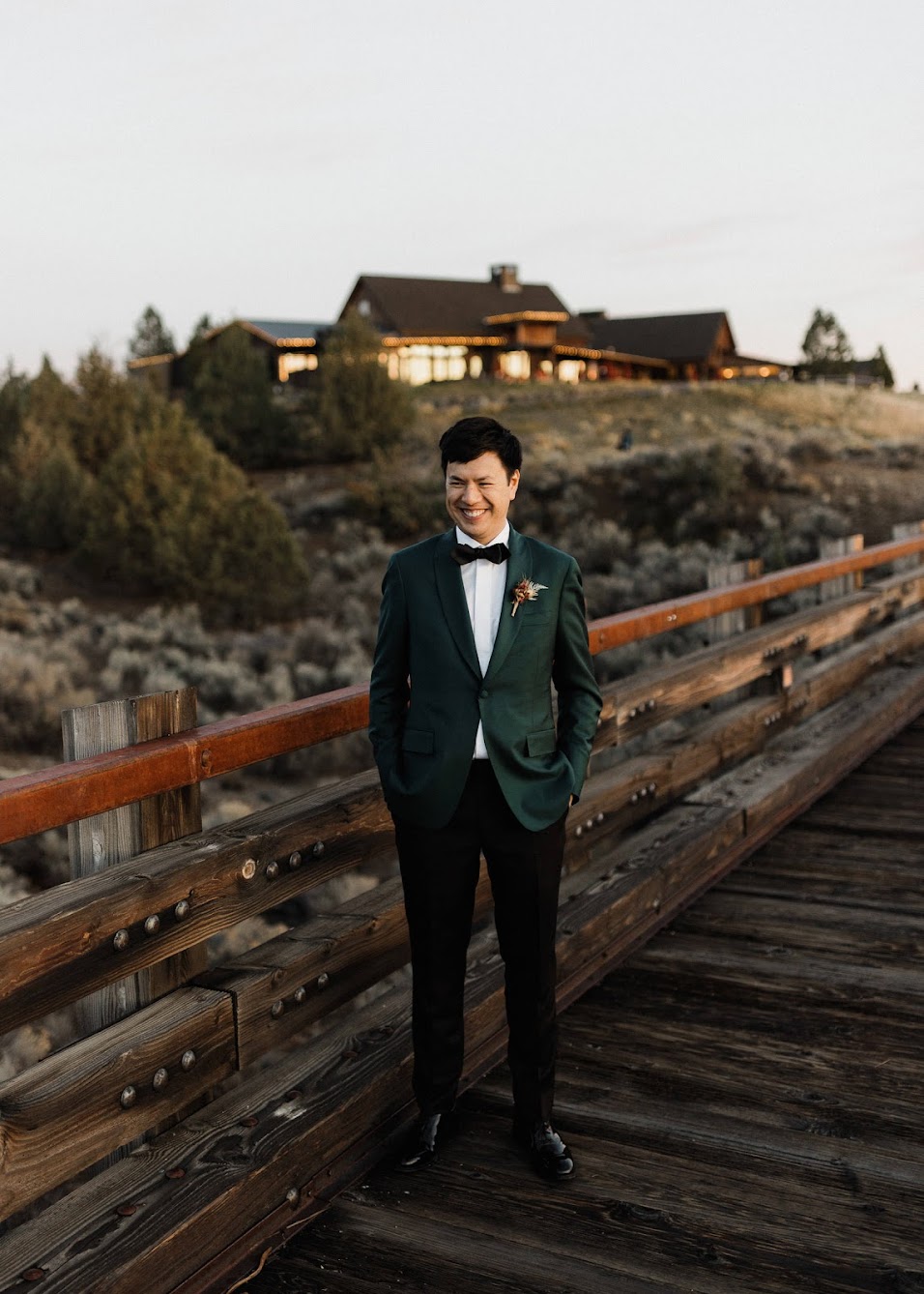 The groom standing alone on the wooden bridge with his hands in his pocket smiling. 