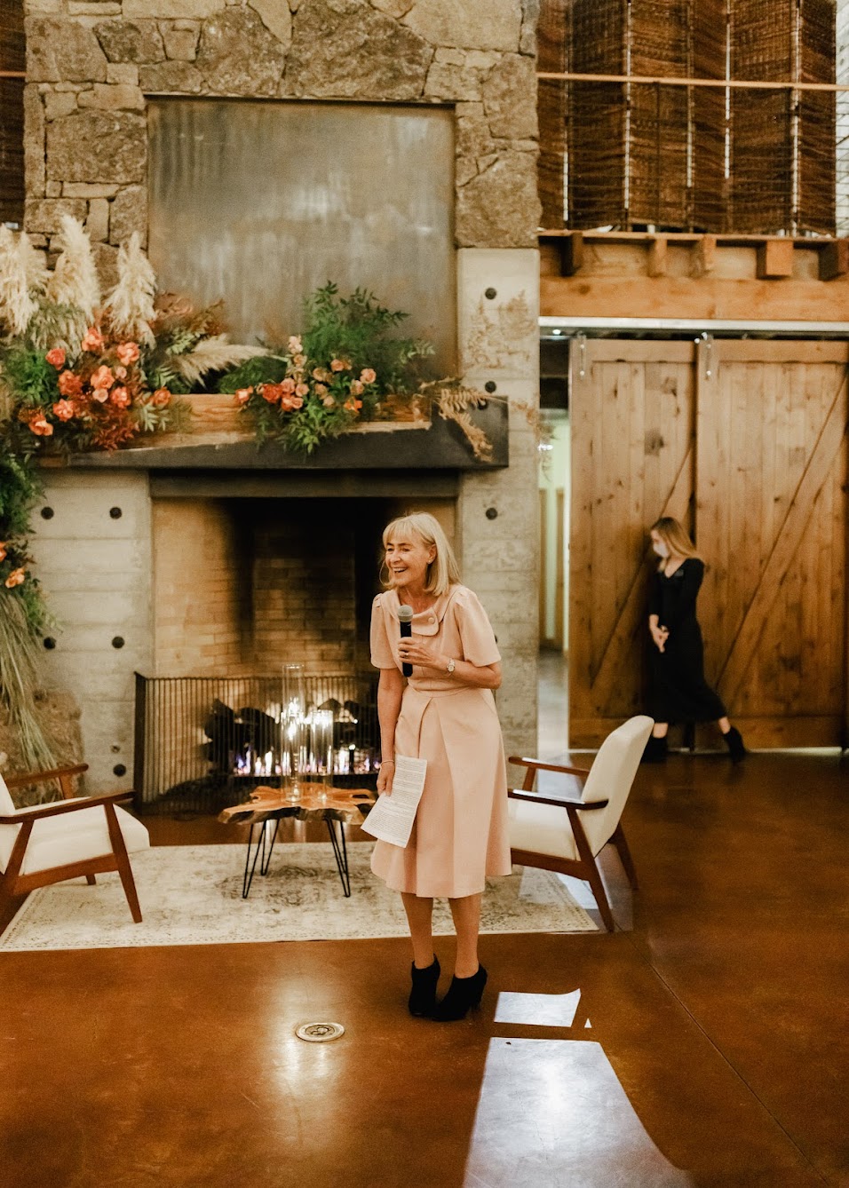 Mother of the bride gives a speech. She stands in front of the lit fireplace with the stunning floral installation on it. She is laughing as she holds the microphone and her paper.