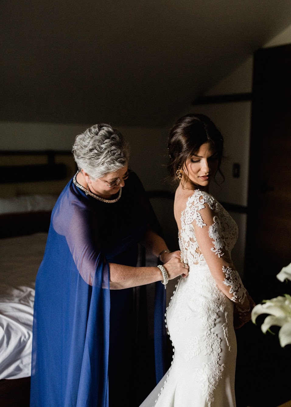 the bride standing in her dress while her mom helps her button it. The dress has buttons that go all the way down to the floor. 