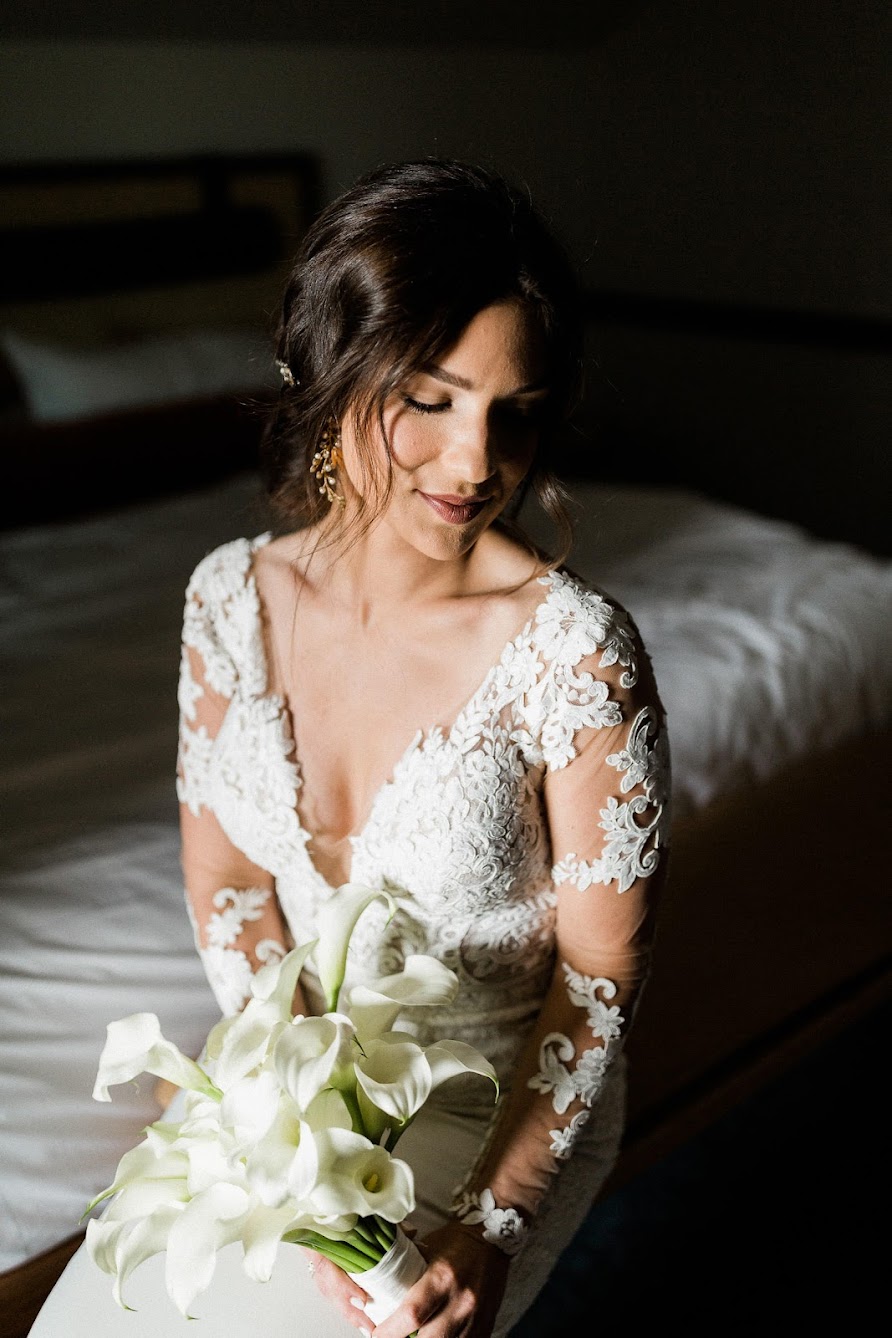 the bride sitting down, the light from the window is hitting her perfectly, lighting up only her and leaving the room dark. She is holding her bridal bouquet.