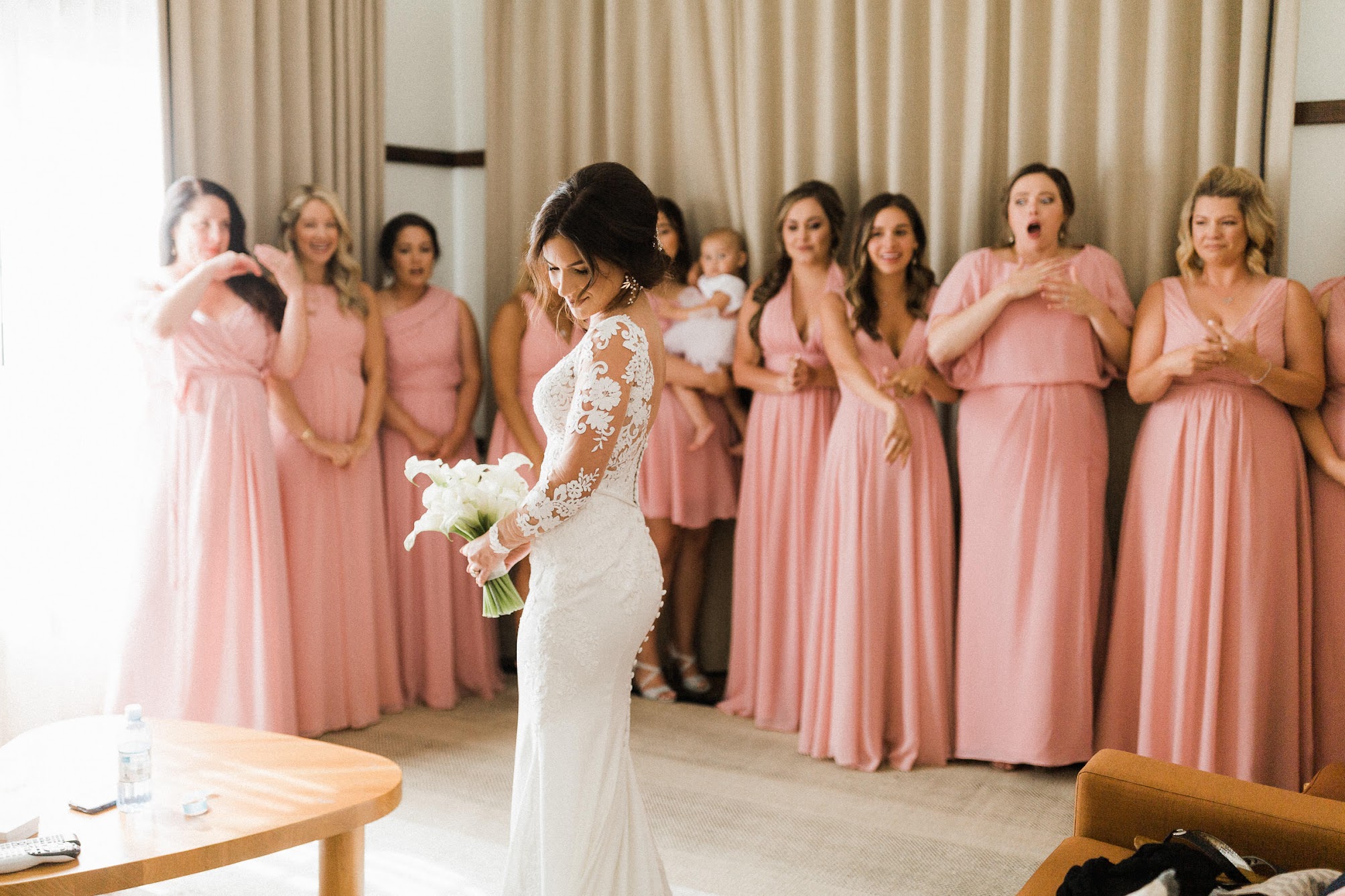 bride showing off her complete look to her bridesmaids. They are all smiling at her.