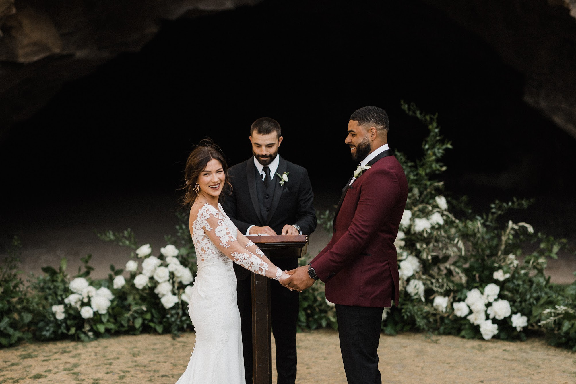 bride and groom holding hands, behind them is their officiant and a stunning line of florals and greenery that stand tall.