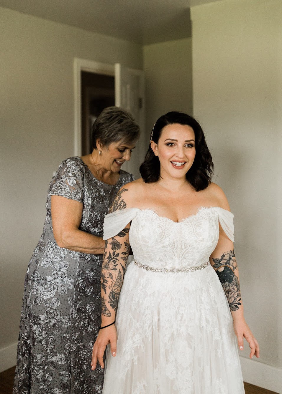 Mother of the bride helping the bride get into her dress. The bride has floral tattoos on both her arms