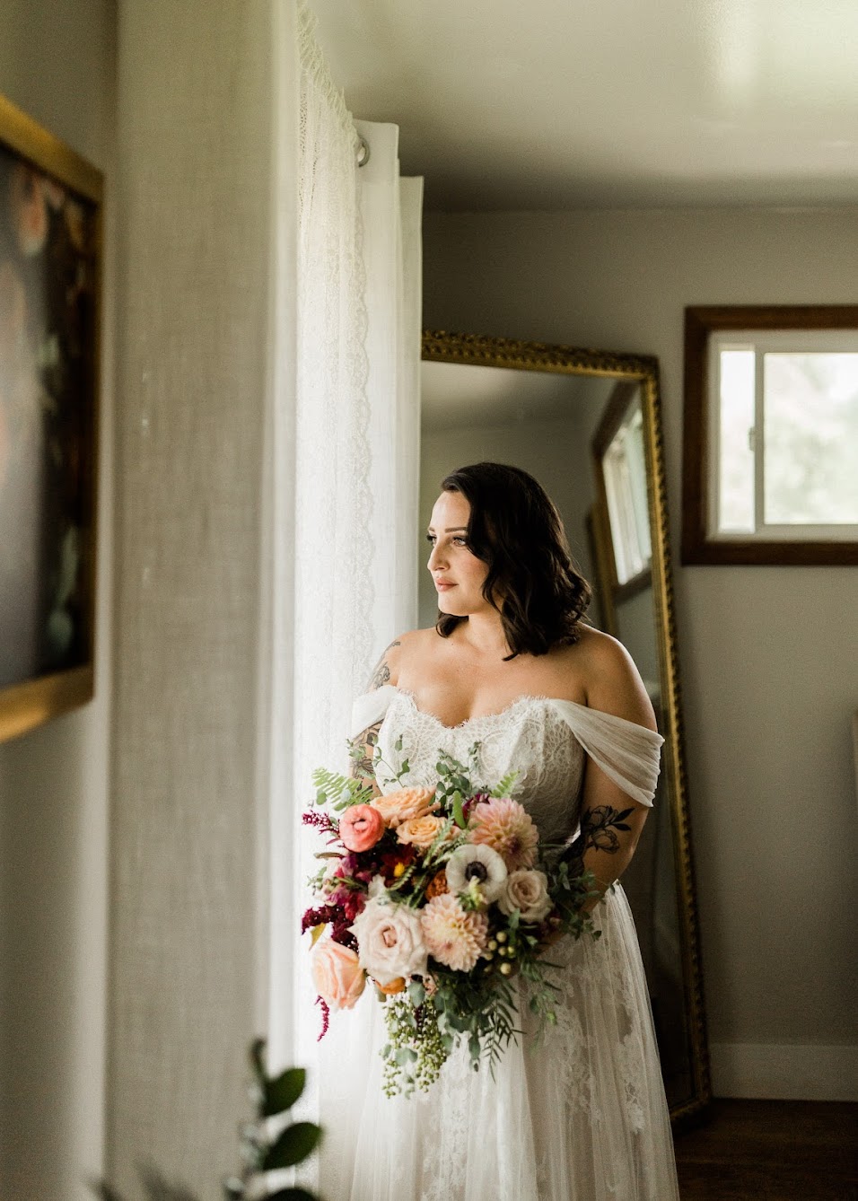 The bride all ready for her big day, staring out of the window while holding her florals wearing her wedding dress.