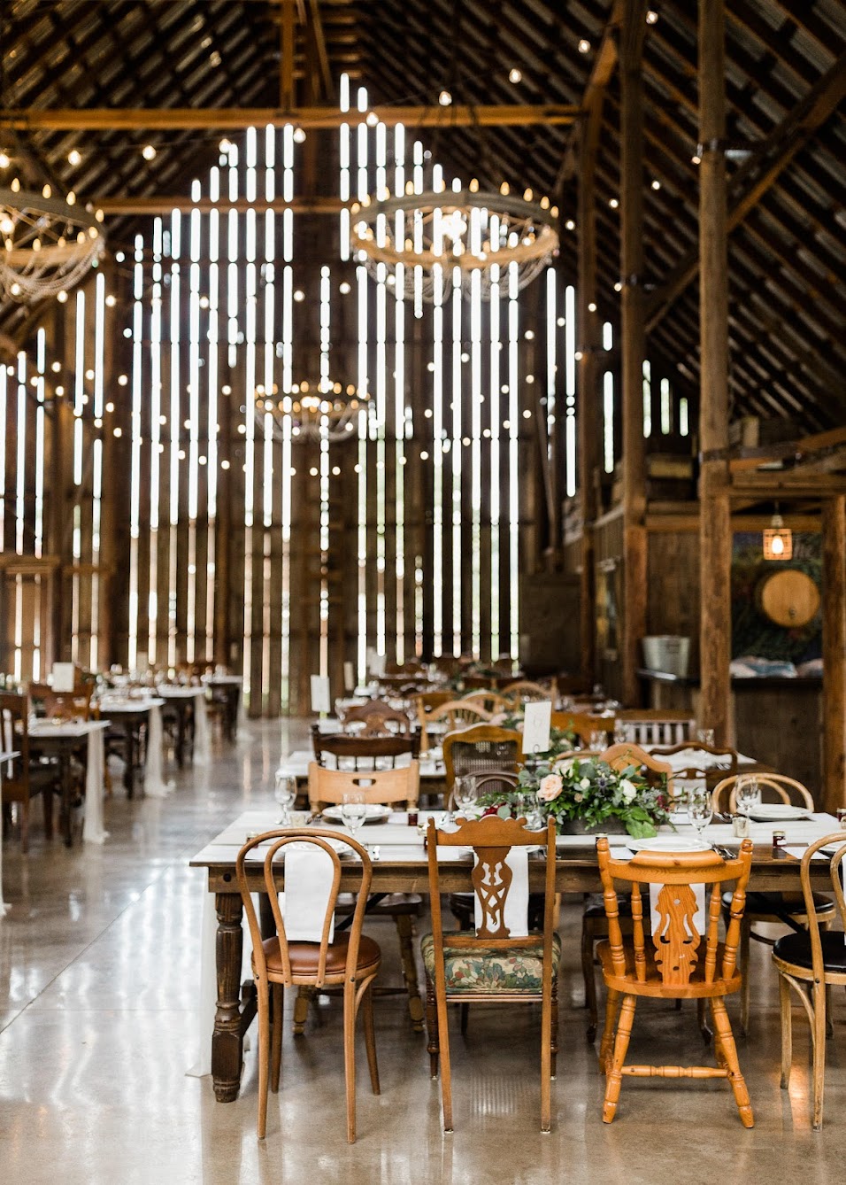 A wedding reception set in a rustic barn, with farm tables and a mix of different wooden chairs.