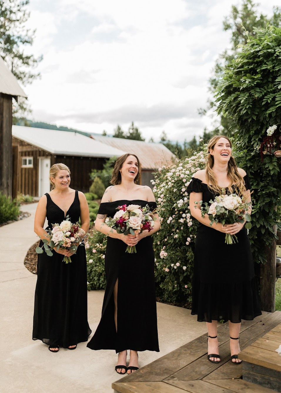 Three bridesmaids wearing black dresses that are all different styles, holding their colorful florals. 