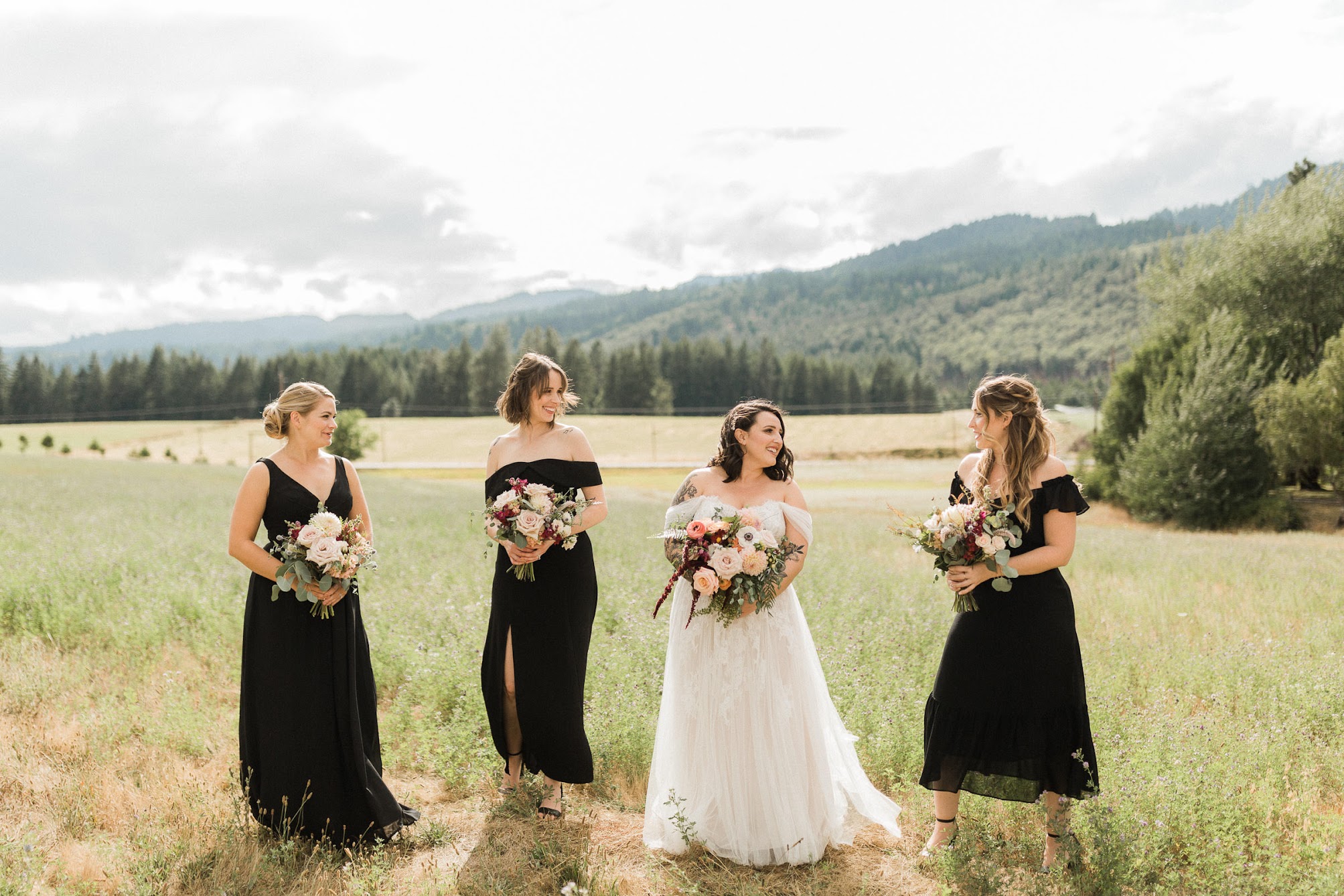 The bride and her three bridesmaids standing apart in a grassy field looking at each other smiling.  