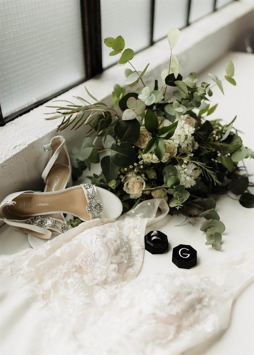 detailed shot of bridal accessories including open-toed wedding heels, floral bouquet, wedding dress, and wedding rings.