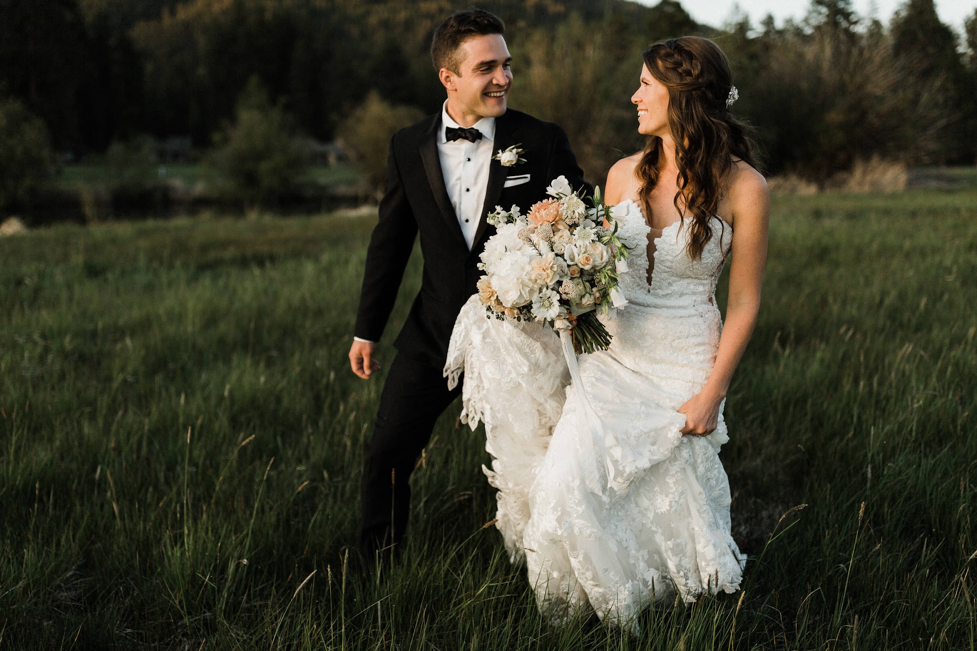 couple walking in tall grass looking each other, the groom is holding the bride's dress 