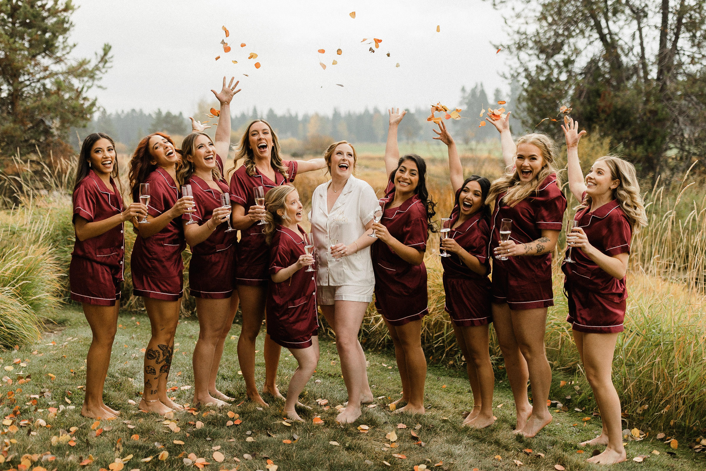 Bride and bridesmaids standing on a lawn, holding champagne glasses, and throwing fall leaves into the air