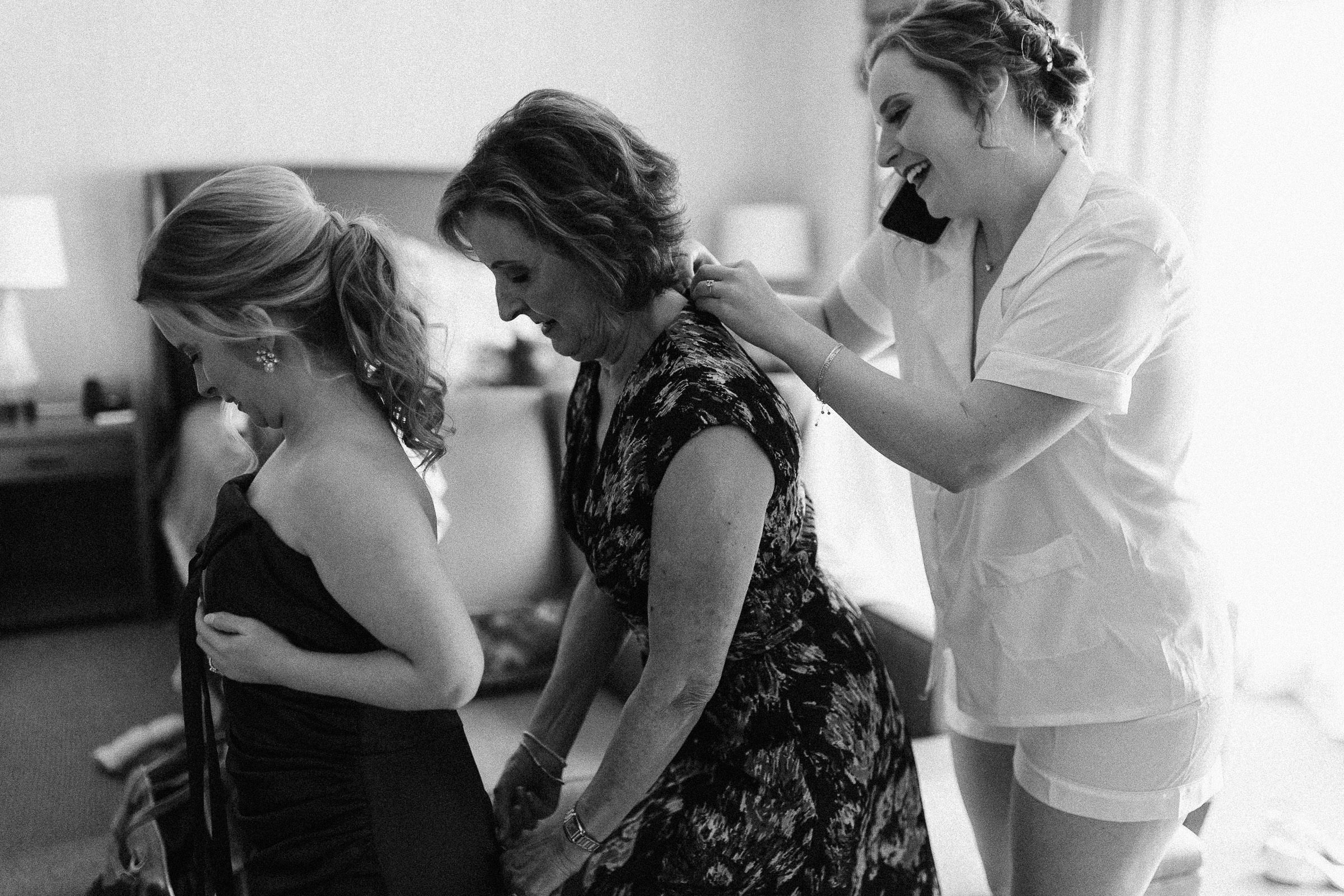 Bride holds smartphone to her ear with her shoulder while buttoning the dress of her mother, who is zipping the dress of her sister