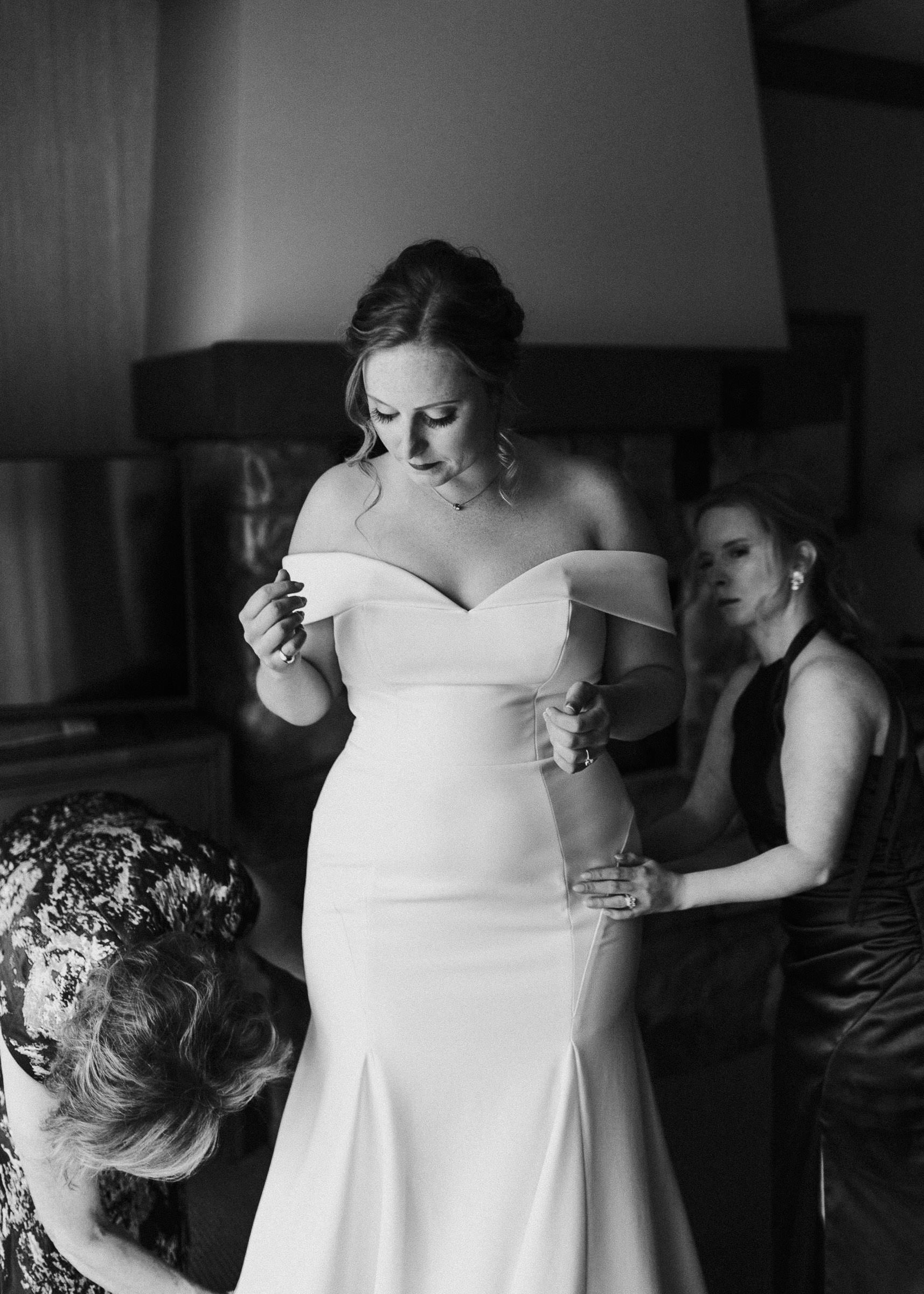Bride looks on as her mom and sister help get her wedding dress on