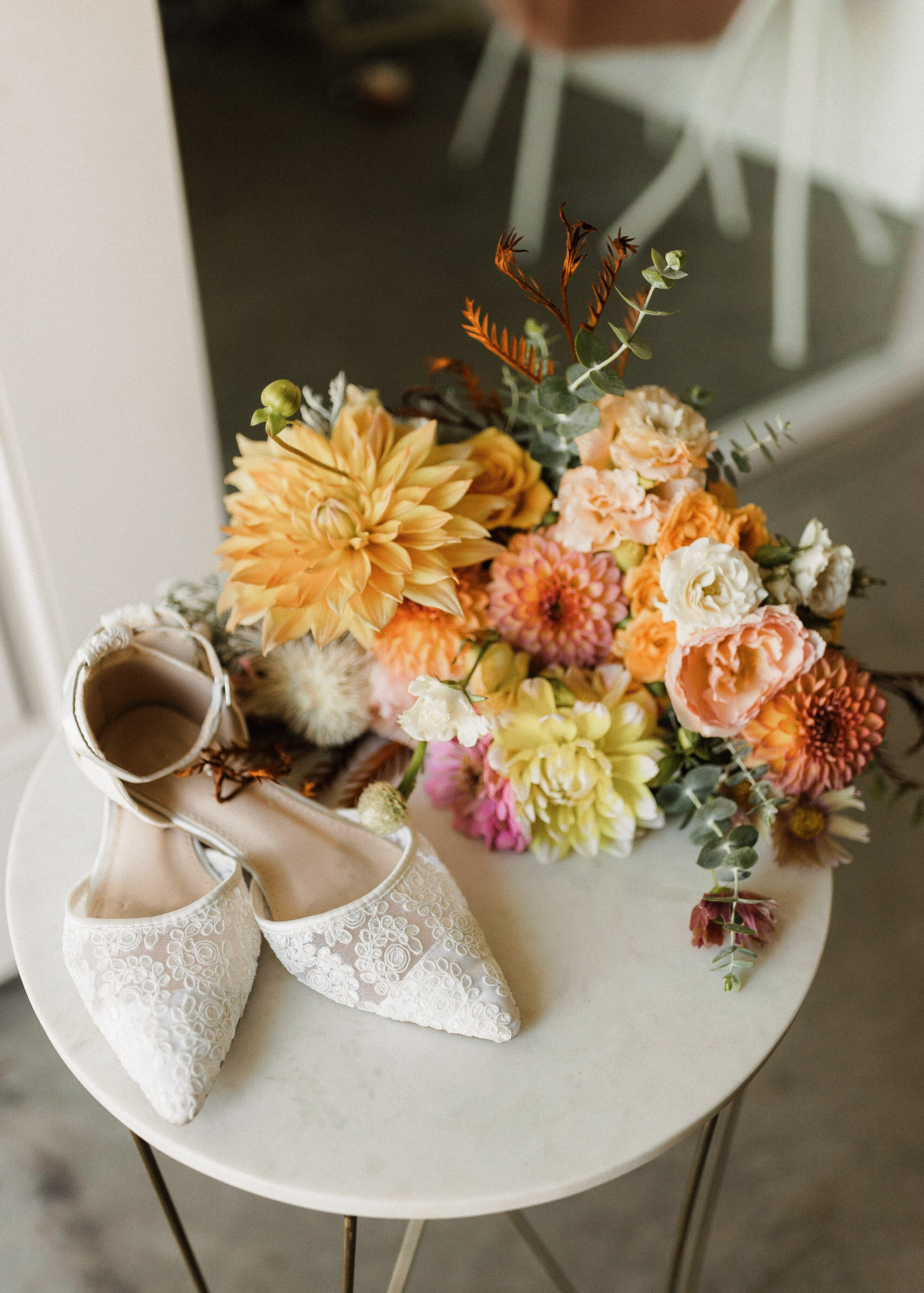 White, lacy wedding shoes rest on a side table with a bridal bouquet in shades of peach, pink, and yellow