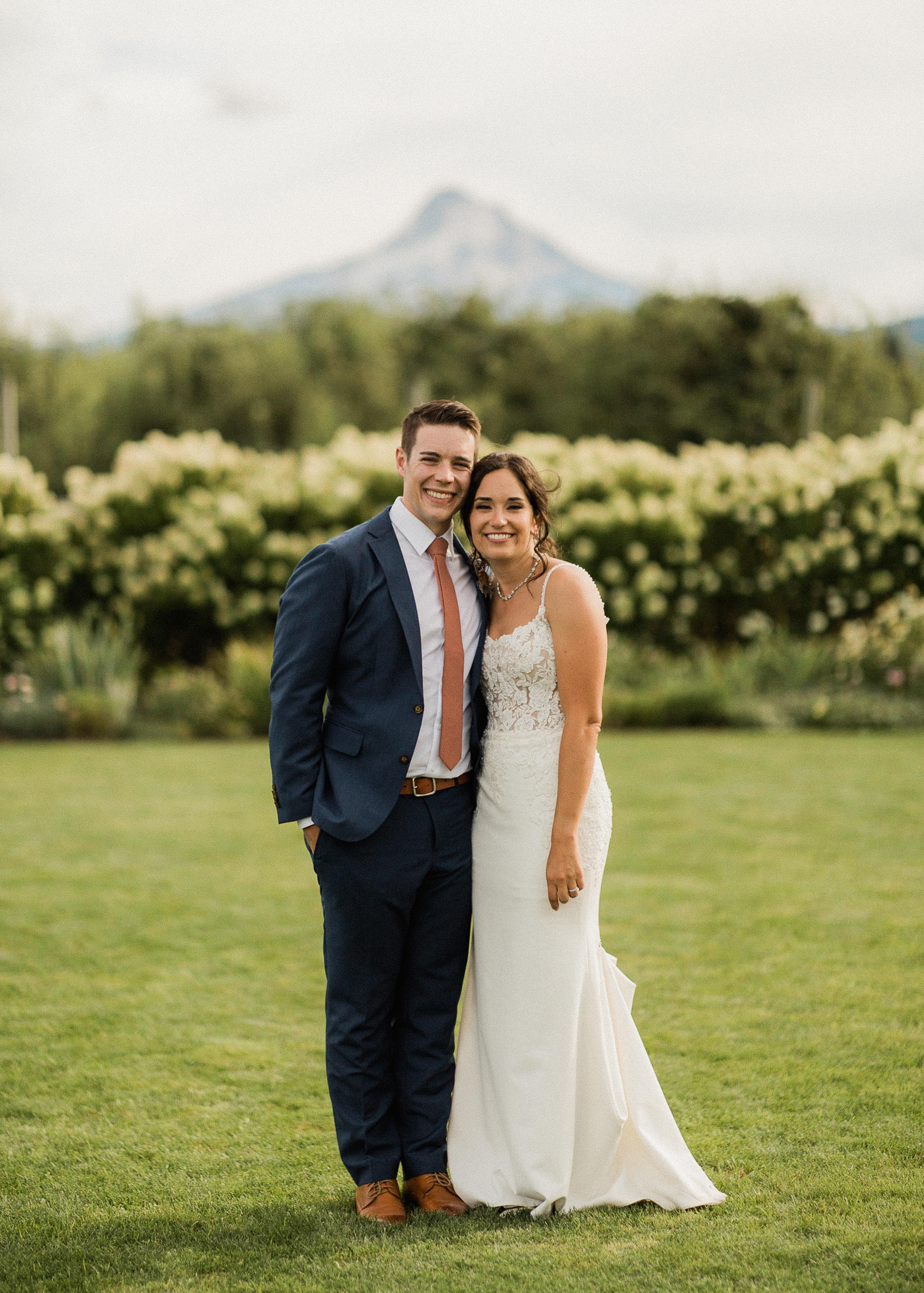 Bride and groom pose in front of an orchard with a view of Mount Hood