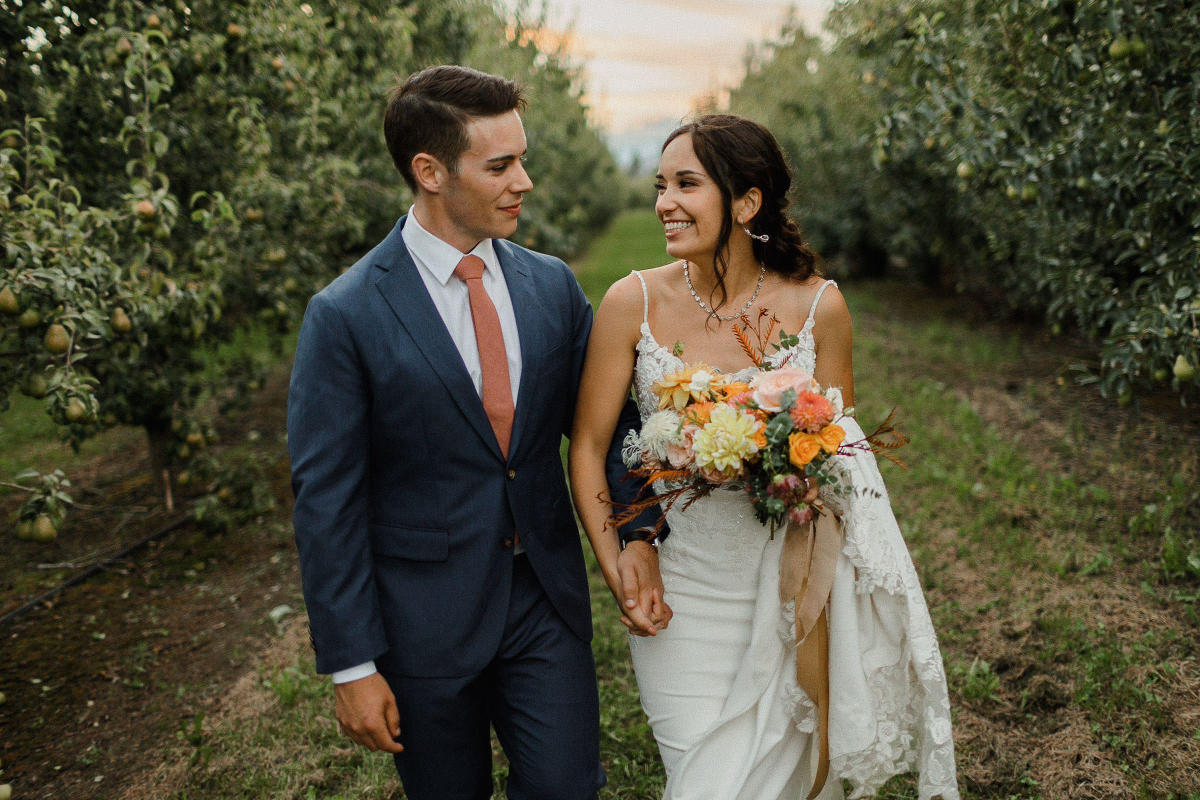 Bride and groom hold hands while walking down an orchard row