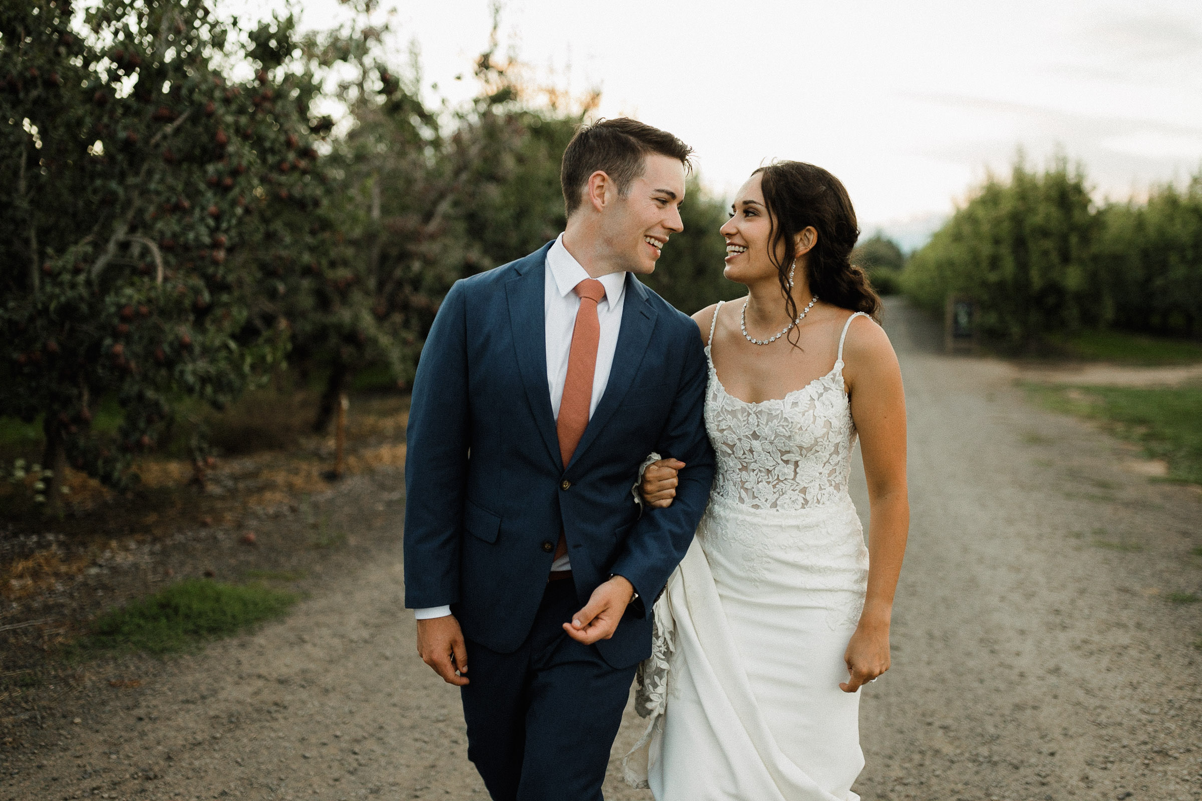 Bride and groom laugh while walking down a gravel road in an orchard